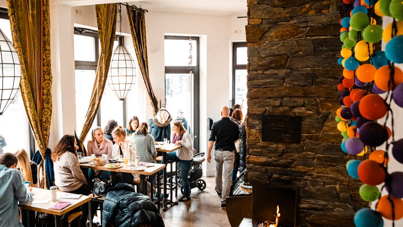 Gem&uuml;tliches Caf&eacute; mit gro&szlig;en Fenstern, Menschen an Tischen, bunte Kugeln an der Wand und ein Kamin. Warme, einladende Atmosph&auml;re., &copy; Stuttgart-Marketing GmbH, Sarah Schmid