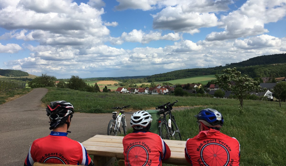 Drei Radfahrer in roten Trikots sitzen auf einer Bank und blicken auf eine ländliche Landschaft mit Feldern und Hügeln unter einem bewölkten Himmel., © Land der 1000 Hügel - Kraichgau-Stromberg