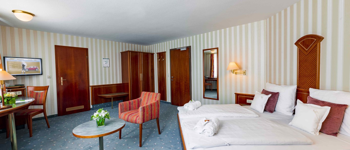 Ein gemütliches Hotelzimmer mit Doppelbett, Tisch, Stuhl und Schrank. Die Wände sind gestreift, der Teppich ist blau. Ein Fernseher hängt an der Wand., © TOMAS