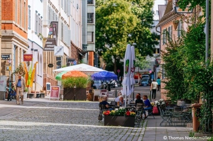 Belebte Straße mit Cafés, bunten Sonnenschirmen und Menschen, die an Tischen sitzen. Gepflasterter Weg, umgeben von Bäumen und Gebäuden., © Stuttgart Marketing GmbH