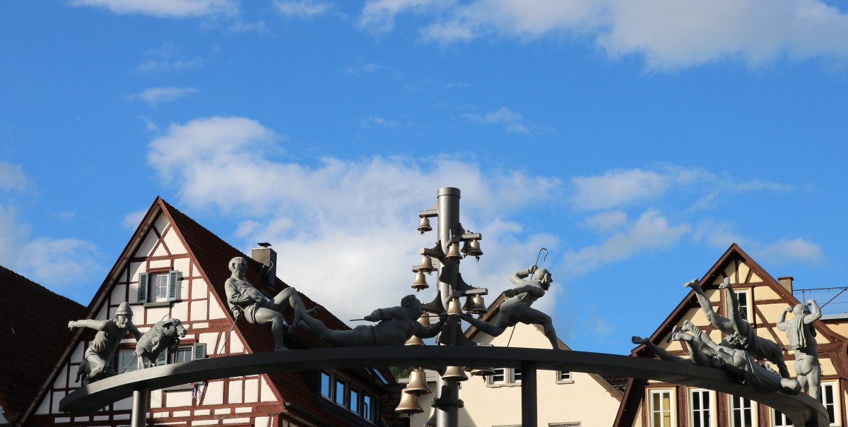 Das Schäferlauf-Denkmal zeigt Figuren und Glocken auf einem Bogen vor Fachwerkhäusern unter blauem Himmel., © Bad Urach Tourismus