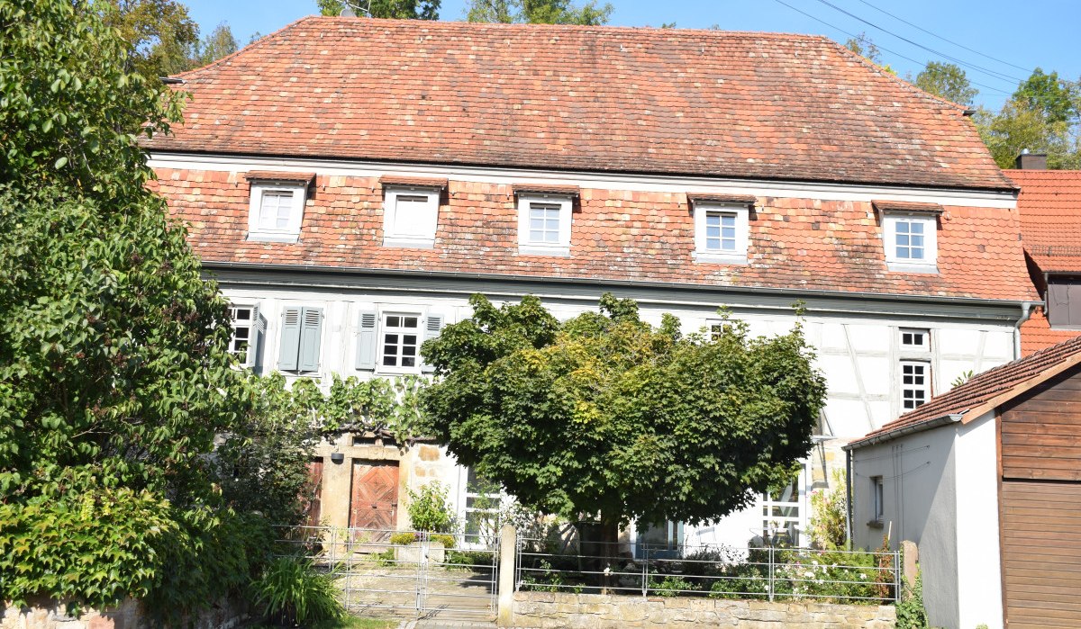 Ein ehemaliges Bauernhaus mit Fachwerk und rotem Ziegeldach, umgeben von Bäumen und Pflanzen. Der Eingangsbereich ist von Grün umrahmt., © Land der 1000 Hügel - Kraichgau-Stromberg Ein ehemaliges Bauernhaus mit Fachwerk und rotem Ziegeldach, umgeben von Bäumen und Pflanzen. Der Eingangsbereich ist von Grün umrahmt., © Land der 1000 Hügel - Kraichgau-Stromberg