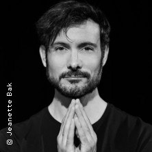 Ein Mann mit Bart und gefalteten Händen blickt in die Kamera. Der Hintergrund ist schwarz. Foto von Jeanette Bak., © links im Bild