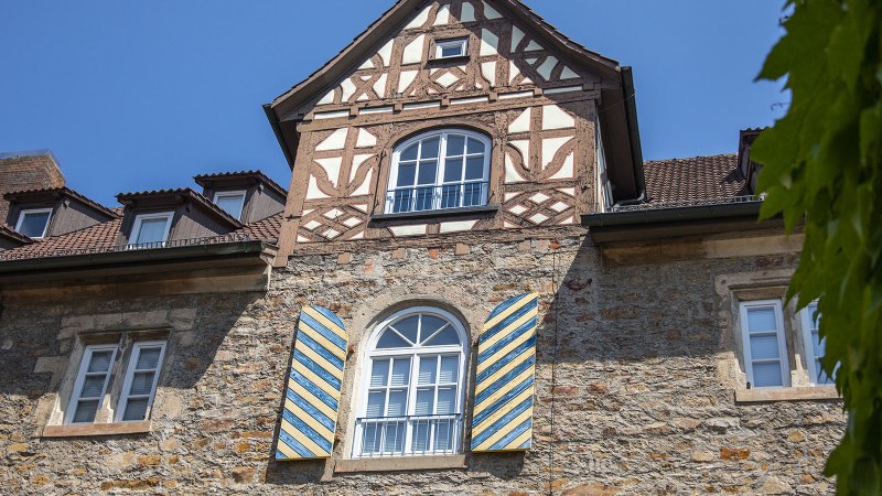 Fachwerkhaus mit blauen Fensterläden, Ziegeldach und grünem Laub im Vordergrund unter blauem Himmel., © Achim Mende Fachwerkhaus mit blauen Fensterläden, Ziegeldach und grünem Laub im Vordergrund unter blauem Himmel., © Achim Mende