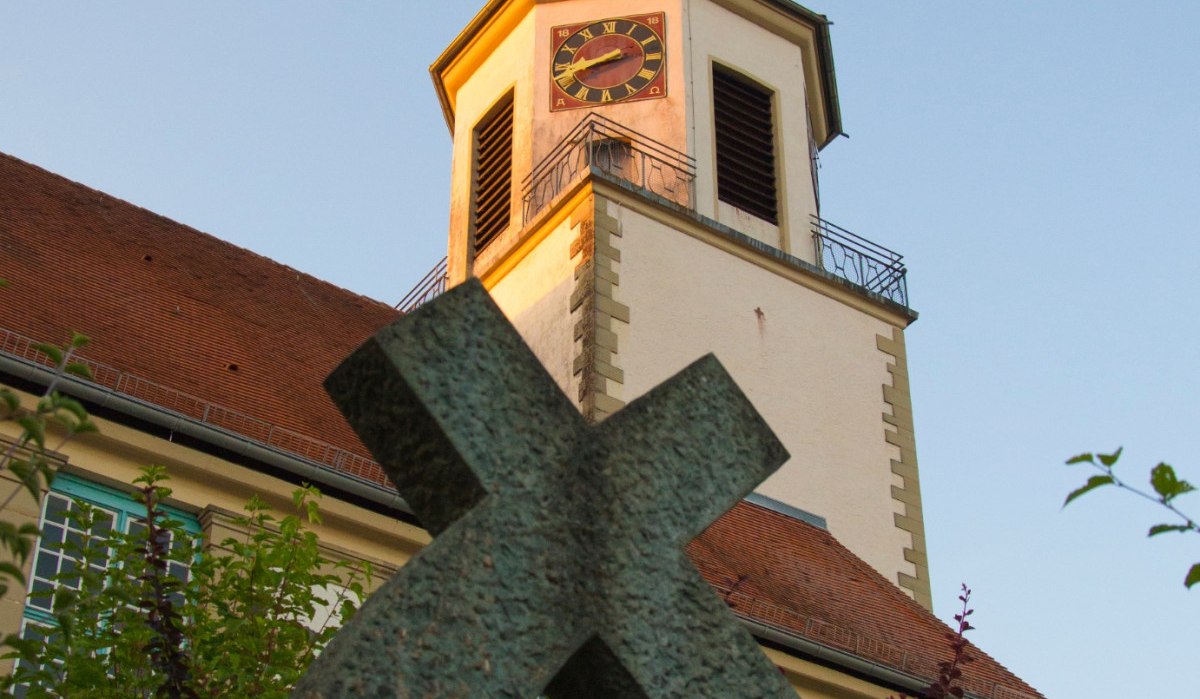 Ein Kirchturm mit Uhr und Wetterhahn ragt in den blauen Himmel. Im Vordergrund steht ein schiefes Kreuz, umgeben von grünen Pflanzen., © Unbekannt Ein Kirchturm mit Uhr und Wetterhahn ragt in den blauen Himmel. Im Vordergrund steht ein schiefes Kreuz, umgeben von grünen Pflanzen., © Unbekannt