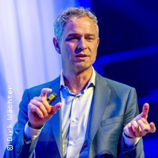 Ein Mann in einem blauen Anzug hält einen Vortrag. Er gestikuliert mit den Händen und hält einen kleinen Gegenstand., © links im Bild Ein Mann in einem blauen Anzug hält einen Vortrag. Er gestikuliert mit den Händen und hält einen kleinen Gegenstand., © links im Bild