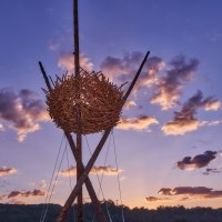 Eine gro&szlig;e Vogelnest-Skulptur aus Holz thront vor einem dramatischen Sonnenuntergang mit violettem Himmel und Wolken., &copy; Natur.Nah. Sch&ouml;nbuch & Heckeng&auml;u