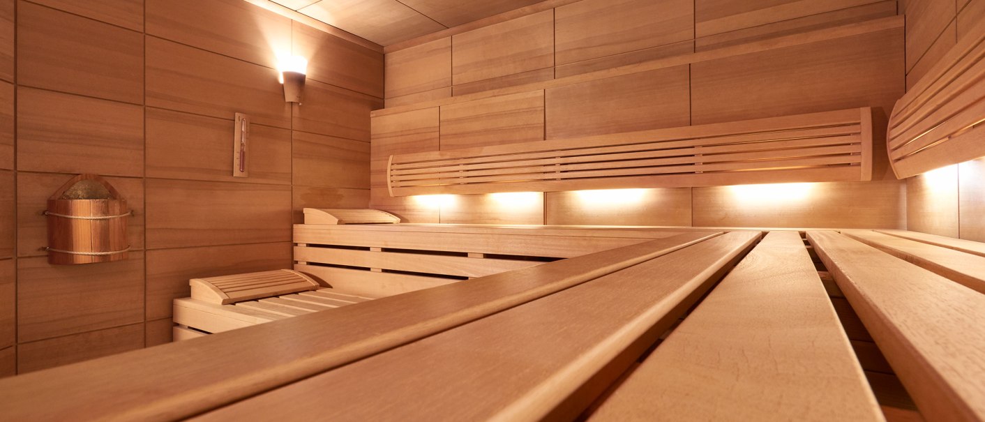Moderne Sauna mit Holzbänken, sanfter Beleuchtung und einem Eimer an der Wand. Die Wände und Decke sind aus hellem Holz., © TOMAS