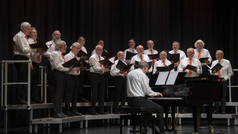 Ein M&auml;nnerchor in wei&szlig;en Hemden und orangefarbenen Krawatten singt auf einer B&uuml;hne. Ein Pianist begleitet sie am Fl&uuml;gel.
