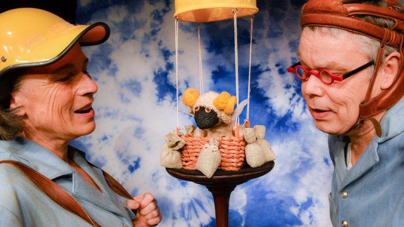 Zwei Personen mit Helmen betrachten ein gestricktes Schaf in einem Mini-Hei&szlig;luftballonkorb vor blau-wei&szlig;em Hintergrund., &copy; FILharmonie Filderstadt