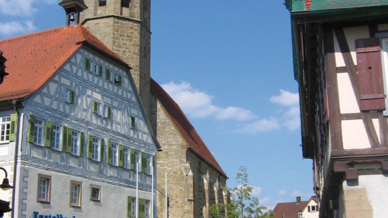 Historische Gebäude in Vaihingen Enzweihingen, darunter ein Kirchturm und Fachwerkhäuser. Ein rotes Auto parkt vor der Enztalbank. Blauer Himmel., © Land der 1000 Hügel - Kraichgau-Stromberg