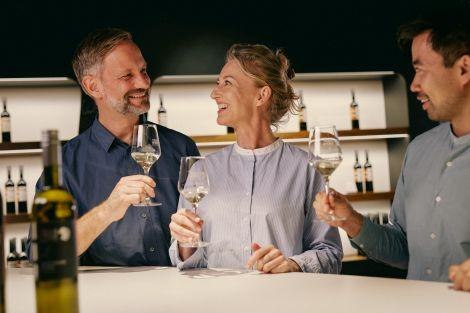 Drei Personen sto&szlig;en mit Weingl&auml;sern an einer modernen Bar an und l&auml;cheln einander zu. Im Hintergrund sind Weinflaschen auf Regalen zu sehen., &copy; Weine in der Tiefe. Foto: Peter Bender