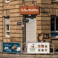 Eckgebäude mit dem Schild 'Ackermanns' an der Fassade. Straßenschilder für Bebelstr. und Schwabstr. sind sichtbar, ebenso mehrere Werbeplakate., © SMG, Sarah Schmid