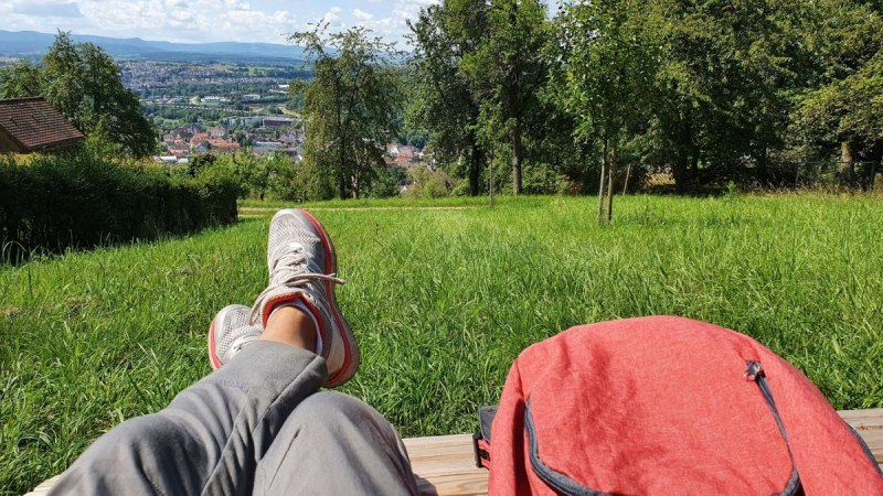 Person entspannt auf einer Bank mit Blick auf eine Stadt in der Ferne, umgeben von gr&uuml;nen Wiesen und B&auml;umen. Ein roter Rucksack liegt daneben.