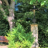 Ein steinernes Kreuz mit Jesusfigur steht neben einer Holzbank unter einem Baum, umgeben von dichtem Grün., © Touristik und Marketing GmbH