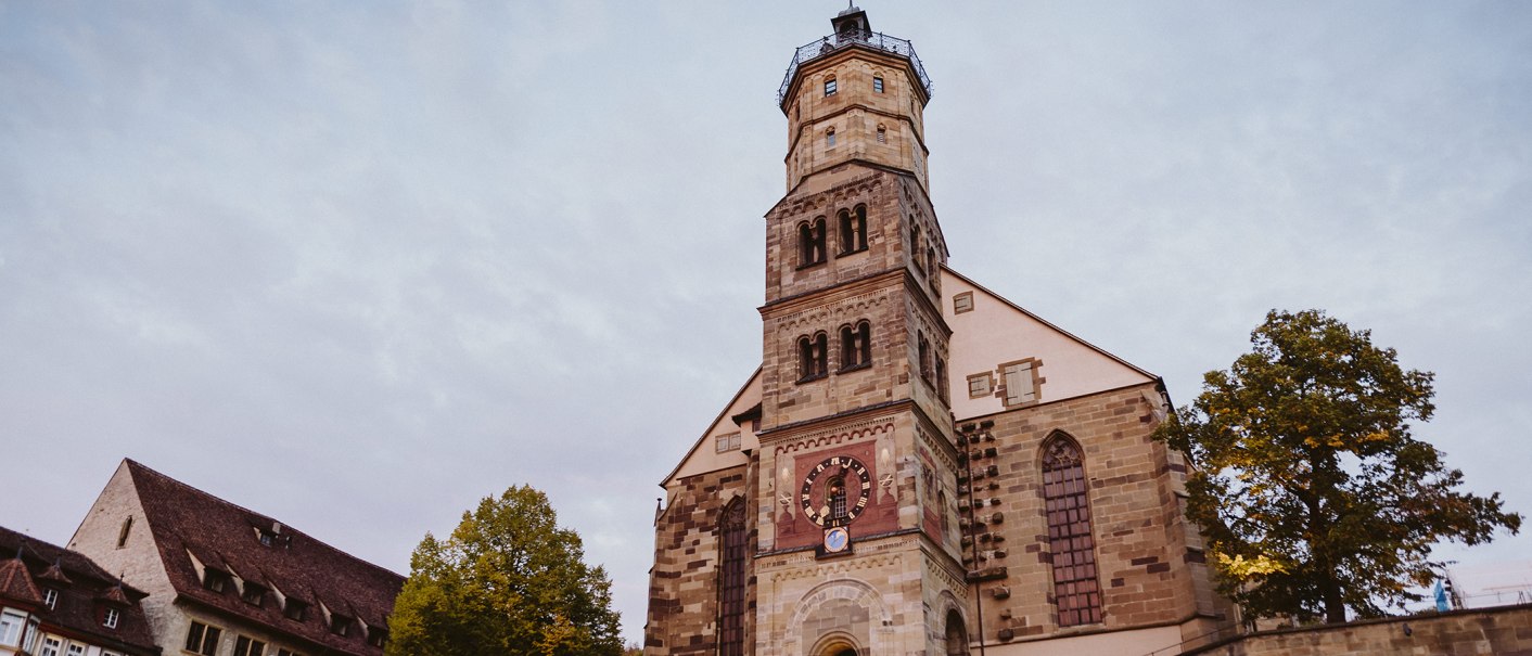 Die Michaelskirche in Schwäbisch Hall mit ihrem markanten Turm und einer Uhr, umgeben von Bäumen, vor einem bewölkten Himmel., © Nico Kurth Die Michaelskirche in Schwäbisch Hall mit ihrem markanten Turm und einer Uhr, umgeben von Bäumen, vor einem bewölkten Himmel., © Nico Kurth