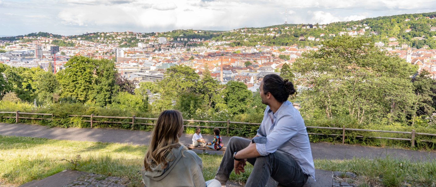 Zwei Personen sitzen auf einer Wiese und schauen auf die Stadt Stuttgart. Im Hintergrund sind Hügel und Gebäude zu sehen, darunter der Fernsehturm., © SMG, Martina Denker