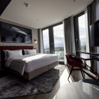 Modernes Eckzimmer mit großem Bett, Schreibtisch, Panoramafenstern und stilvollem Dekor. Ein Bild eines Autos hängt an der Wand., © Radisson Hotel Group Modernes Eckzimmer mit großem Bett, Schreibtisch, Panoramafenstern und stilvollem Dekor. Ein Bild eines Autos hängt an der Wand., © Radisson Hotel Group