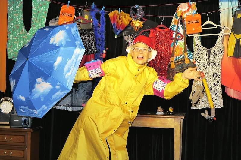 Eine Person in einem gelben Regenmantel und rotem Hut posiert mit einem blauen Regenschirm vor einer bunten Kulisse aus Kleidung und Accessoires., &copy; Galli Theater Backnang
