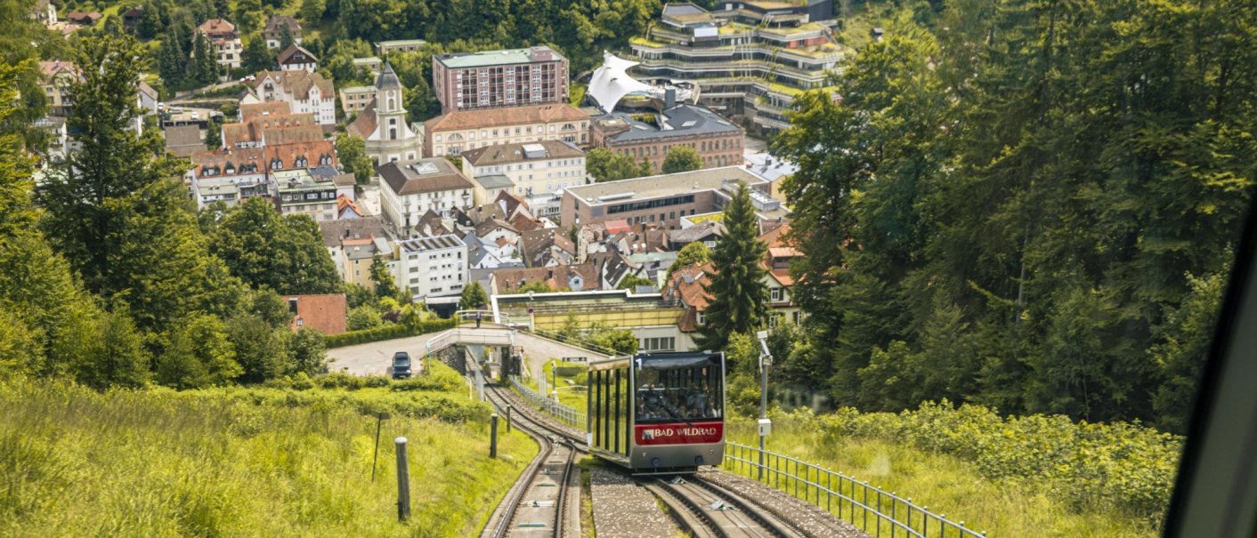 Die Sommerbergbahn fährt durch grüne Landschaft auf Bad Wildbad zu. Im Hintergrund sind Gebäude und Bäume zu sehen., © Stuttgart-Marketing GmbH, Sarah Schmid