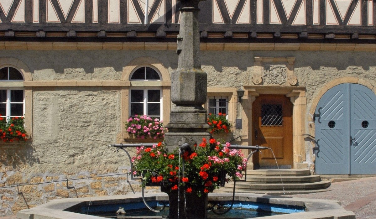 Ein steinerner Brunnen mit Blumen steht vor einem historischen Fachwerkhaus mit blauen Fensterläden und einem Holztor., © Natur.Nah. Schönbuch & Heckengäu Ein steinerner Brunnen mit Blumen steht vor einem historischen Fachwerkhaus mit blauen Fensterläden und einem Holztor., © Natur.Nah. Schönbuch & Heckengäu