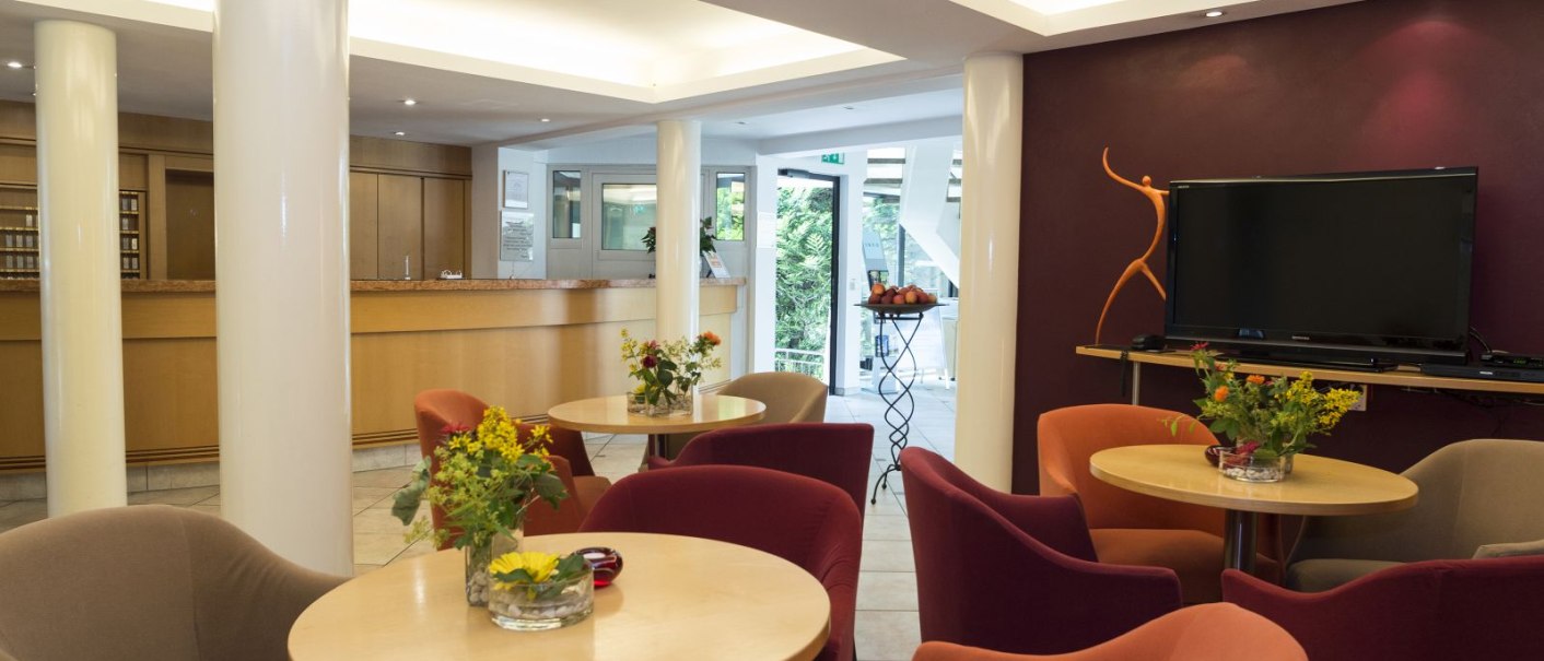 Moderne Hotel-Lobby mit gemütlichen Sesseln, Tischen mit Blumen, einem großen Fernseher und einer Rezeption im Hintergrund., © Hotel Gloria GmbH Restaurant Möhringer Hexle Moderne Hotel-Lobby mit gemütlichen Sesseln, Tischen mit Blumen, einem großen Fernseher und einer Rezeption im Hintergrund., © Hotel Gloria GmbH Restaurant Möhringer Hexle