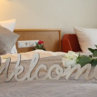 Ein Bett mit Kissen, einer weißen Rose und einem 'Welcome'-Schild. Im Hintergrund eine Steckdose und ein roter Sessel., © TOMAS