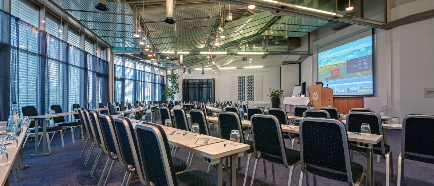 Moderner Tagungsraum mit Stuhlreihen, Tischen und einem großen Bildschirm. Die Decke ist mit Beleuchtung und Lüftung ausgestattet., © City Hotel Reutlingen