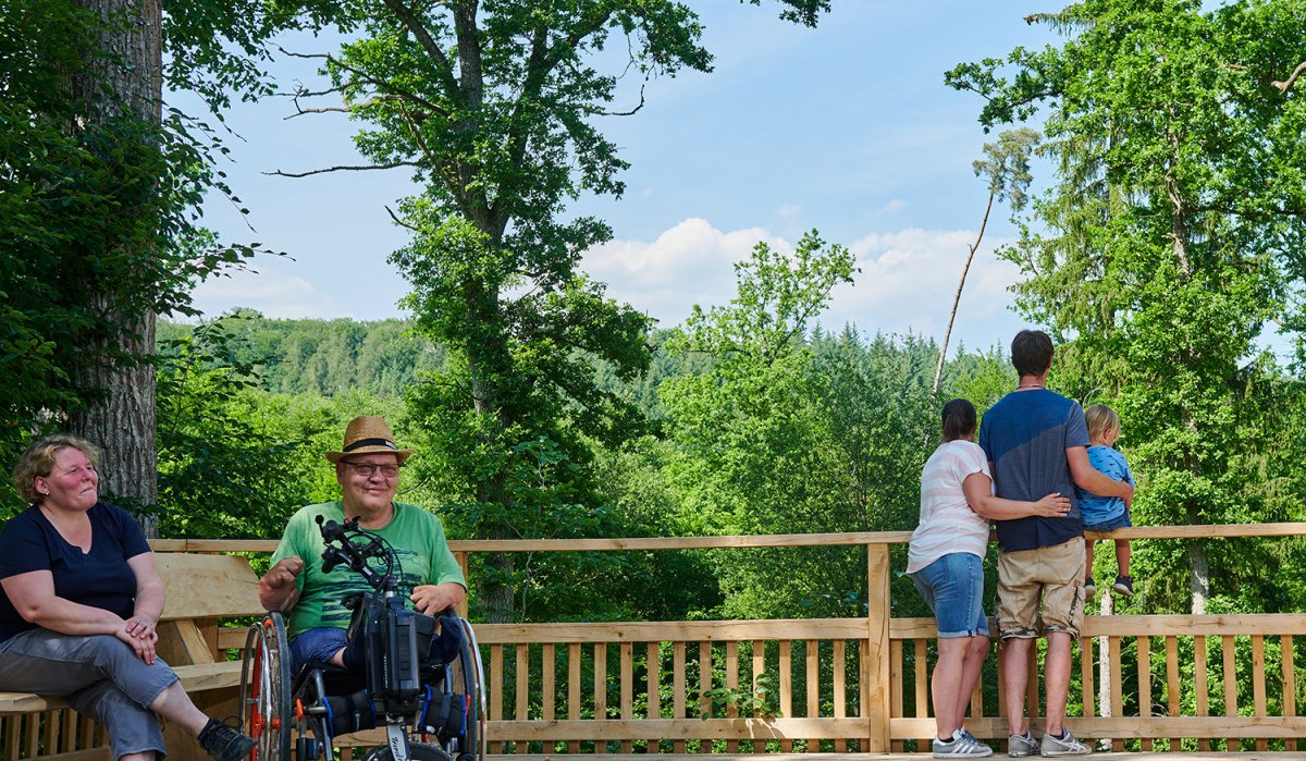 Menschen genie&szlig;en die Aussicht von einer Holzplattform im Wald. Eine Person im Rollstuhl l&auml;chelt, w&auml;hrend eine Familie die Landschaft betrachtet.