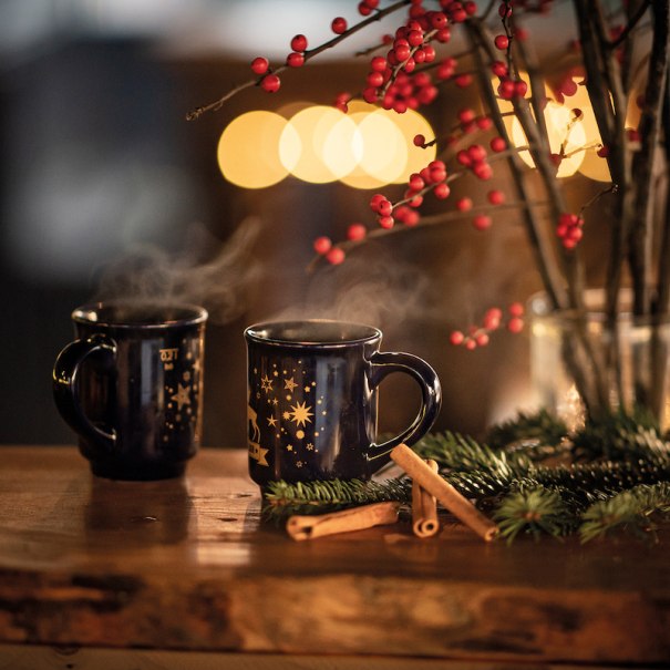 Zwei dampfende Tassen mit Weihnachtsmotiven stehen auf einem Tisch, umgeben von Zimtstangen, Tannenzweigen und roten Beeren. Im Hintergrund leuchten Lichter., © Mercedes-Benz AG Zwei dampfende Tassen mit Weihnachtsmotiven stehen auf einem Tisch, umgeben von Zimtstangen, Tannenzweigen und roten Beeren. Im Hintergrund leuchten Lichter., © Mercedes-Benz AG