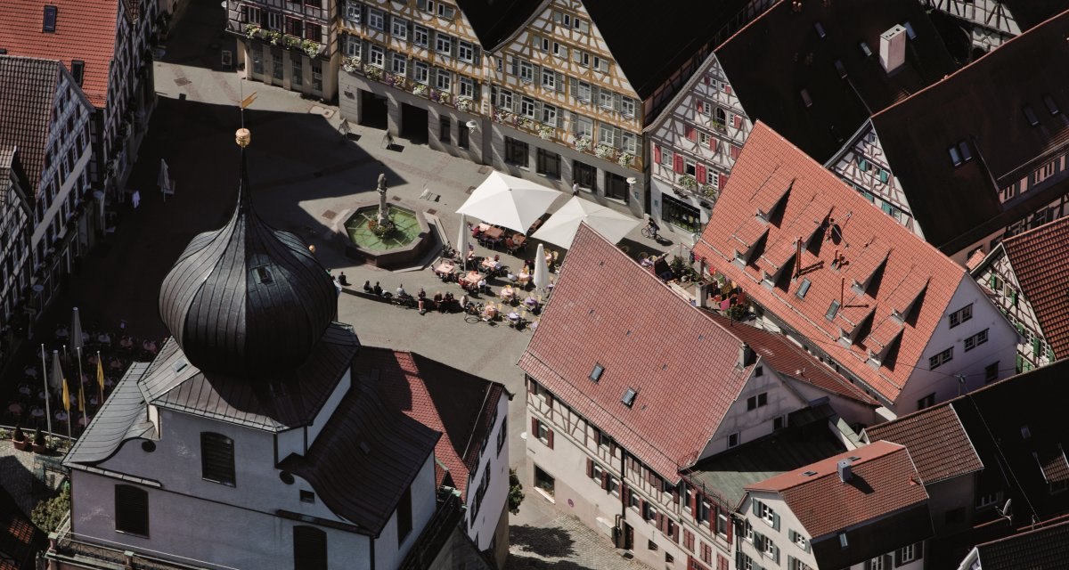 Luftaufnahme eines Marktplatzes mit Fachwerkh&auml;usern, einem Brunnen und einem Geb&auml;ude mit Zwiebelturm. Menschen sitzen unter Sonnenschirmen., &copy; Regio Marketing GmbH