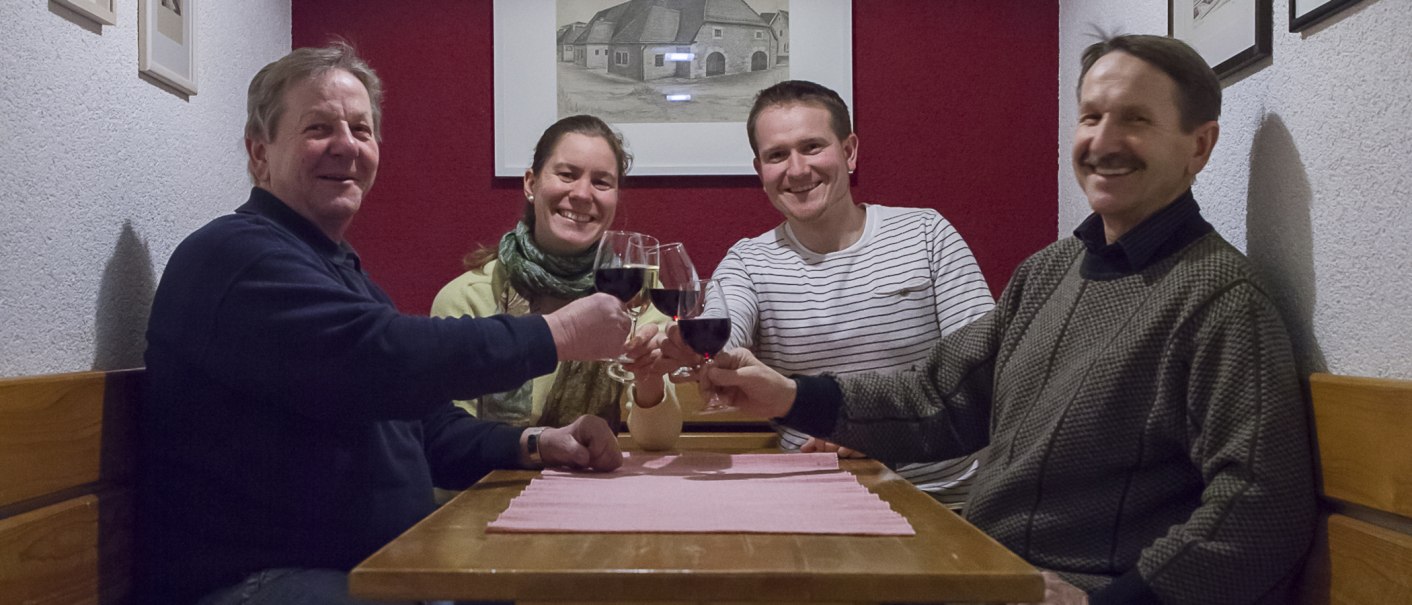 Vier Personen sitzen an einem Tisch und stoßen mit Weingläsern an. Sie lächeln und wirken fröhlich. Im Hintergrund hängt ein Bild an der Wand., © 70469R!