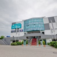 Das Hotel Le Village zeigt eine moderne Fassade mit Glasfront und Treppe. Vor dem Gebäude wehen Fahnen, der Himmel ist bewölkt., © Hotel Le Village