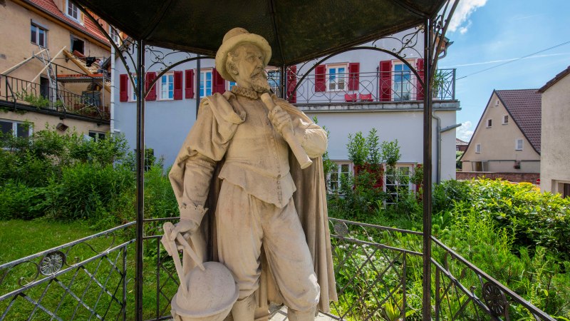 Steinstatue eines Mannes mit Hut und Umhang, der ein Instrument hält. Sie steht unter einem Pavillon in einem grünen Garten, umgeben von Häusern., © Stuttgart-Marketing GmbH, Achim Mende