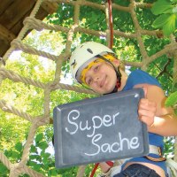Person im Kletterpark hält eine Tafel mit der Aufschrift 'Super Sache' und trägt einen Helm. Im Hintergrund sind Bäume und ein Kletternetz zu sehen., © Stuttgart-Marketing GmbH