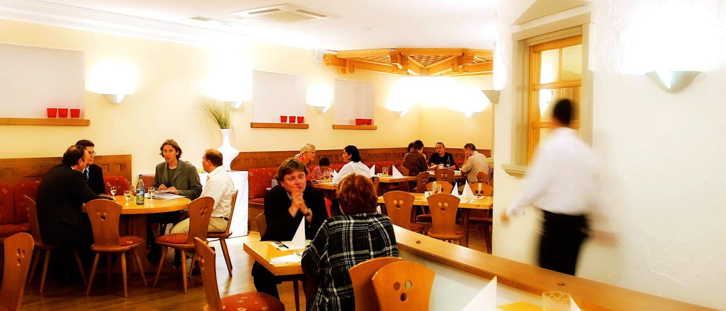 Gemütliches Restaurant mit Holzmöbeln, mehreren Gästen an Tischen und einem Kellner in Bewegung. Warme Beleuchtung und dekorative Elemente an den Wänden., © SMG Gemütliches Restaurant mit Holzmöbeln, mehreren Gästen an Tischen und einem Kellner in Bewegung. Warme Beleuchtung und dekorative Elemente an den Wänden., © SMG