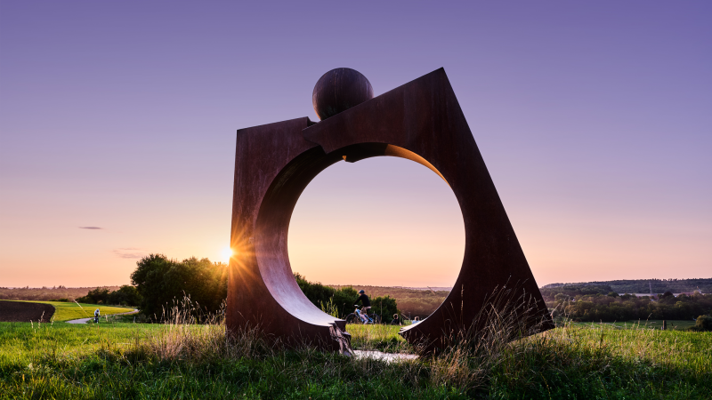 Eine gro&szlig;e Metallskulptur steht auf einem Feld, w&auml;hrend die Sonne am Horizont untergeht. Menschen spazieren und radeln im Hintergrund., &copy; Andreas Sporn