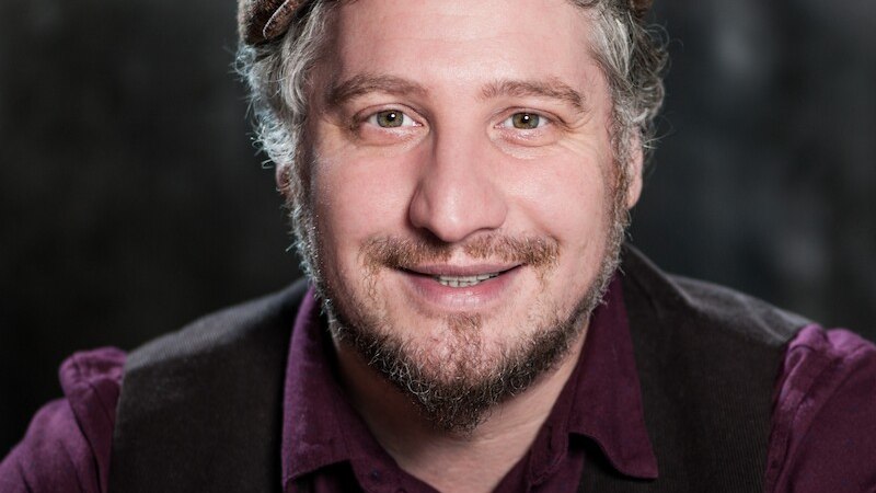 Ein Mann mit Schiebermütze und Bart lächelt in die Kamera. Er trägt ein lila Hemd und eine dunkle Weste. Der Hintergrund ist dunkel und unscharf., © Mauerwerk GmbH Ein Mann mit Schiebermütze und Bart lächelt in die Kamera. Er trägt ein lila Hemd und eine dunkle Weste. Der Hintergrund ist dunkel und unscharf., © Mauerwerk GmbH