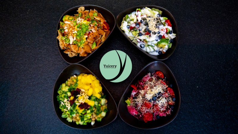 Vier Schalen mit bunten Salaten und Zutaten, darunter Pasta, Obst und Gemüse, auf schwarzem Hintergrund. Logo "Yucery" in der Mitte., © Yuícery, Stuttgart