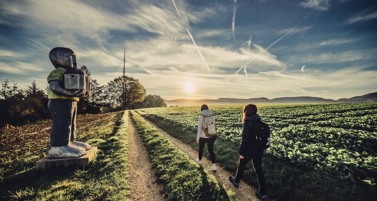 Zwei Personen wandern bei Sonnenaufgang auf einem Feldweg. Links steht eine große Skulptur. Der Himmel ist klar mit einigen Wolkenstreifen., © Landratsamt Ostalbkreis Zwei Personen wandern bei Sonnenaufgang auf einem Feldweg. Links steht eine große Skulptur. Der Himmel ist klar mit einigen Wolkenstreifen., © Landratsamt Ostalbkreis