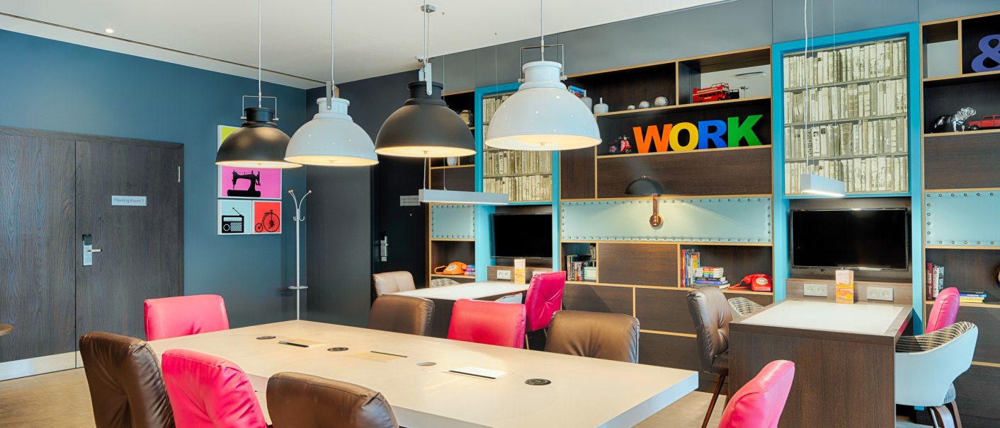 Moderner Arbeitsbereich mit bunten Stühlen, großen Tischen und Regalen. Helle Lampen hängen von der Decke, im Hintergrund steht das Wort 'WORK' in bunten Buchstaben., © Premier Inn Germany