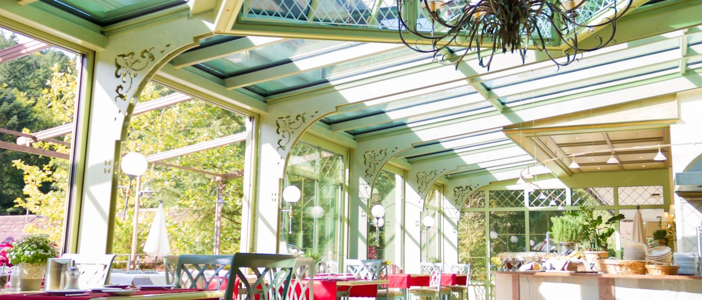 Ein heller Wintergarten mit Glasdach, dekoriert mit Pflanzen und roten Tischdecken. Ein Kronleuchter hängt von der Decke., © TOMAS