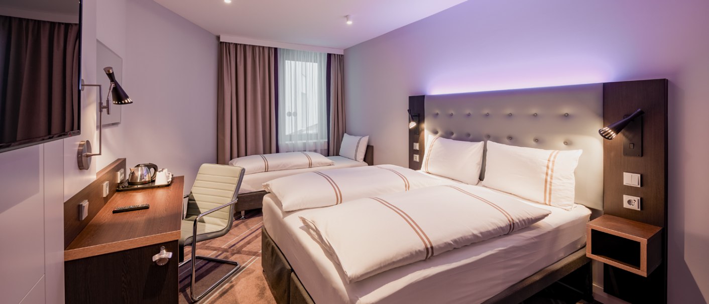 Modernes Dreibettzimmer mit zwei Einzelbetten, Schreibtisch, Fernseher und stilvoller Beleuchtung. Helle Farben und gemütliche Atmosphäre., © Premier Inn