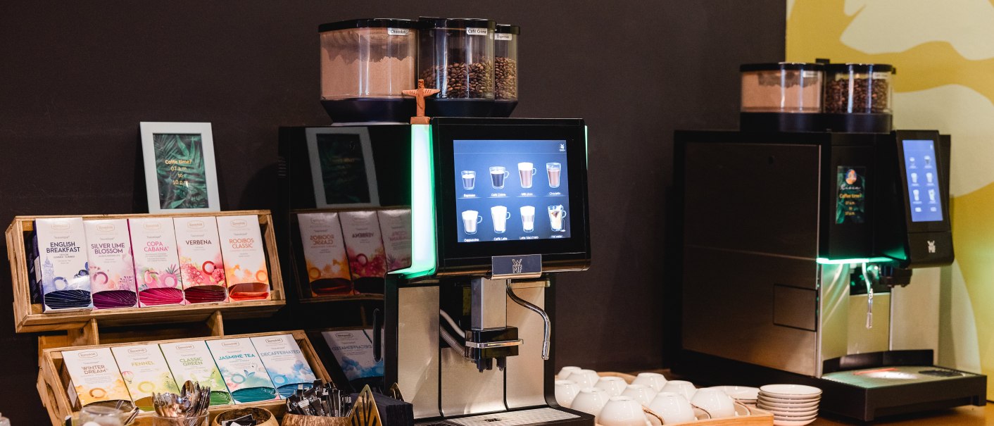 Zwei Kaffeemaschinen mit Touchscreen, Teesortiment und Gebäck auf einem Tisch. Tassen und Löffel sind bereitgestellt., © Rioca Stuttgart Posto 4 GmbH