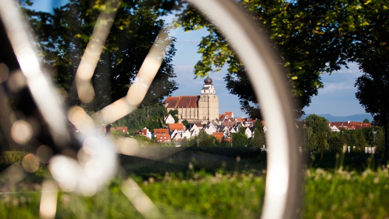 Ein Fahrrad im Vordergrund, durch dessen Speichen eine Kirche in einer Stadt zu sehen ist, umgeben von Bäumen und grüner Landschaft., © Natur.Nah. Schönbuch & Heckengäu Ein Fahrrad im Vordergrund, durch dessen Speichen eine Kirche in einer Stadt zu sehen ist, umgeben von Bäumen und grüner Landschaft., © Natur.Nah. Schönbuch & Heckengäu
