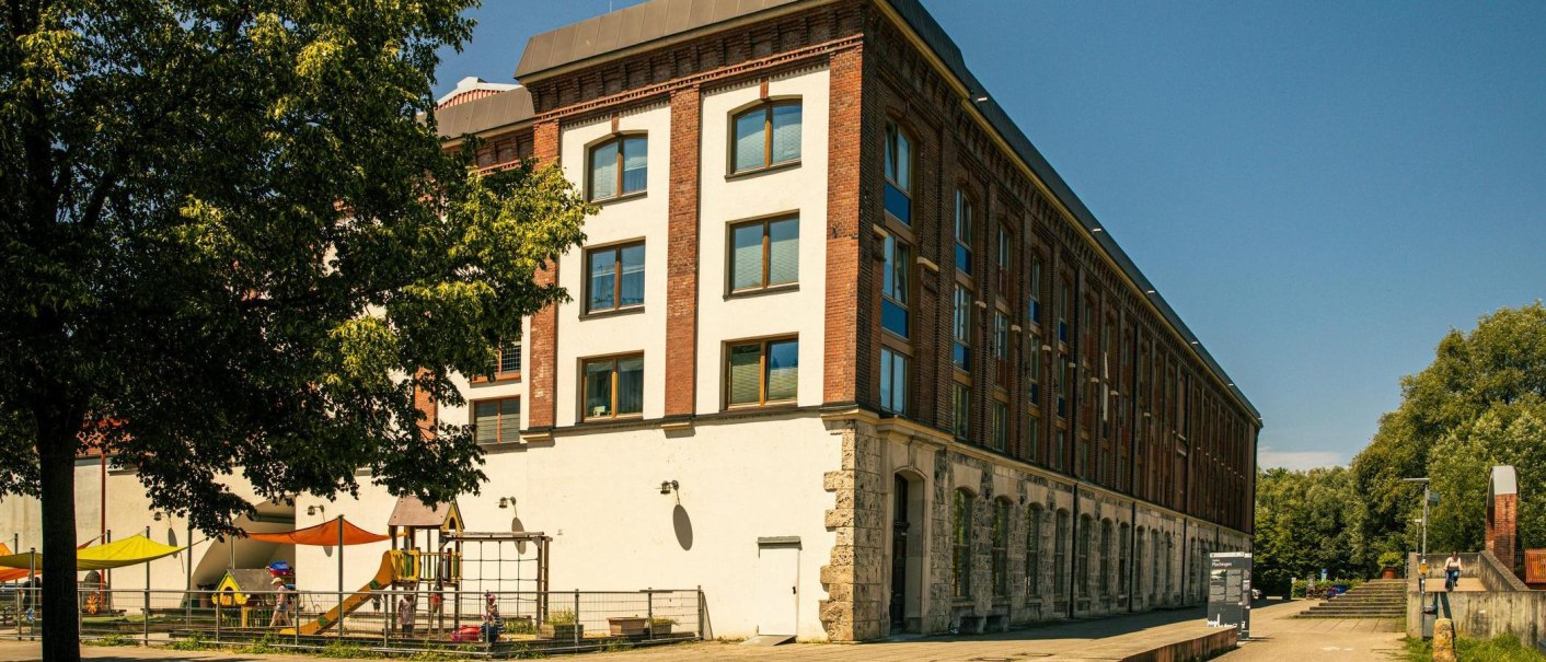 Ein altes Backsteingebäude mit modernen Fenstern, daneben ein Spielplatz mit bunten Sonnensegeln. Bäume im Vordergrund und blauer Himmel., © Stuttgart-Marketing GmbH, Sarah Schmid Ein altes Backsteingebäude mit modernen Fenstern, daneben ein Spielplatz mit bunten Sonnensegeln. Bäume im Vordergrund und blauer Himmel., © Stuttgart-Marketing GmbH, Sarah Schmid