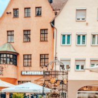 Der Hans-im-Glück-Brunnen vor historischen Gebäuden. Im Vordergrund sind Sonnenschirme und Figuren zu sehen. Die Häuser haben rote Dächer und viele Fenster., © SMG, takemyhearteverywhere