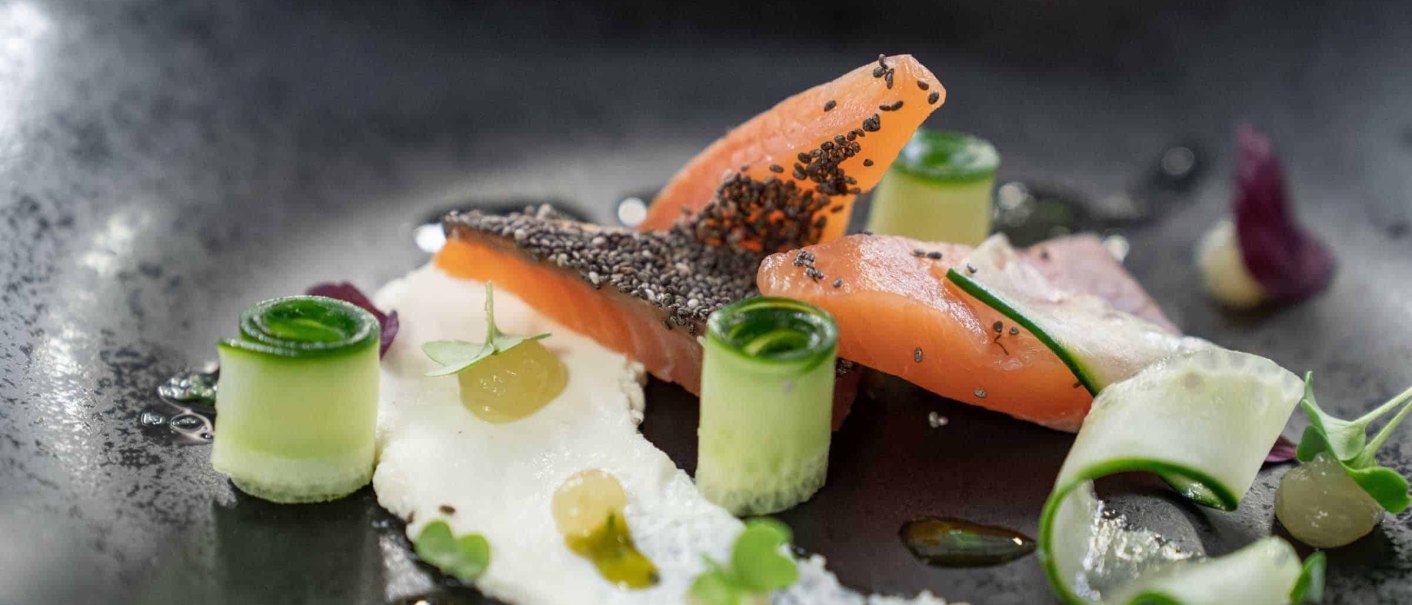 Gourmetgericht mit Lachs, Chiasamen, Gurkenröllchen und Creme auf schwarzem Teller kunstvoll angerichtet., © Flairhotel Vier Jahreszeiten