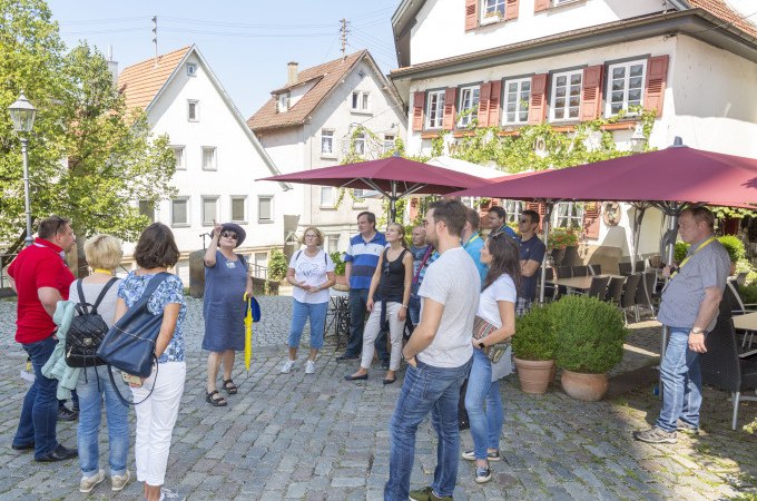 Gruppe bei Stadtführung auf gepflastertem Platz vor historischen Gebäuden mit roten Fensterläden und Sonnenschirmen., © Stadt Nürtingen
