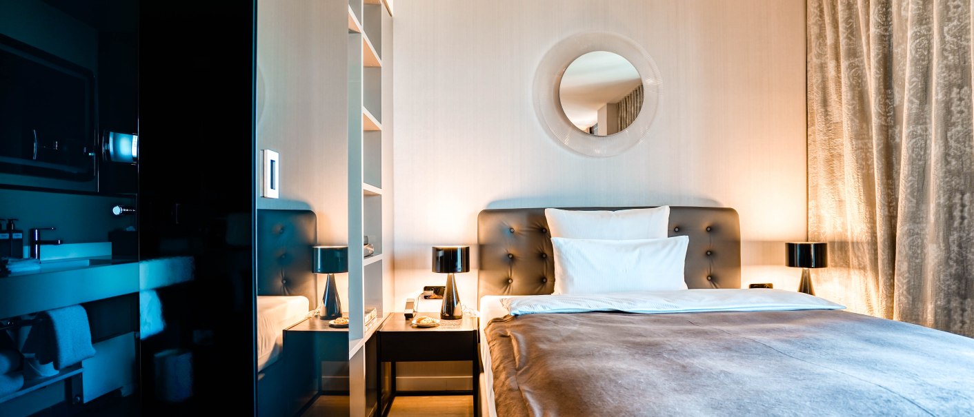 Modernes Hotelzimmer mit elegantem Doppelbett, runden Spiegel über dem Kopfteil, Nachttischen mit Lampen und Vorhängen an der Seite., © CLOUD N°7 APARTMENTS GmbH